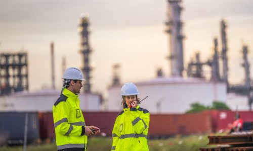 Two engineers in high-visibility safety gear and helmets conduct a field inspection at an industrial plant during sunrise, holding a tablet and measuring tools with soft pastel light background.